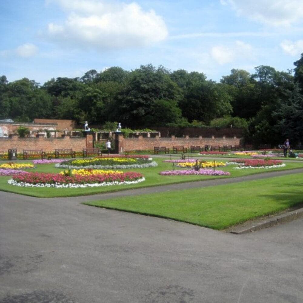 Beautiful garden with colorful flower beds, paved paths, green lawns, benches, and a brick wall in the background. - Home Instead