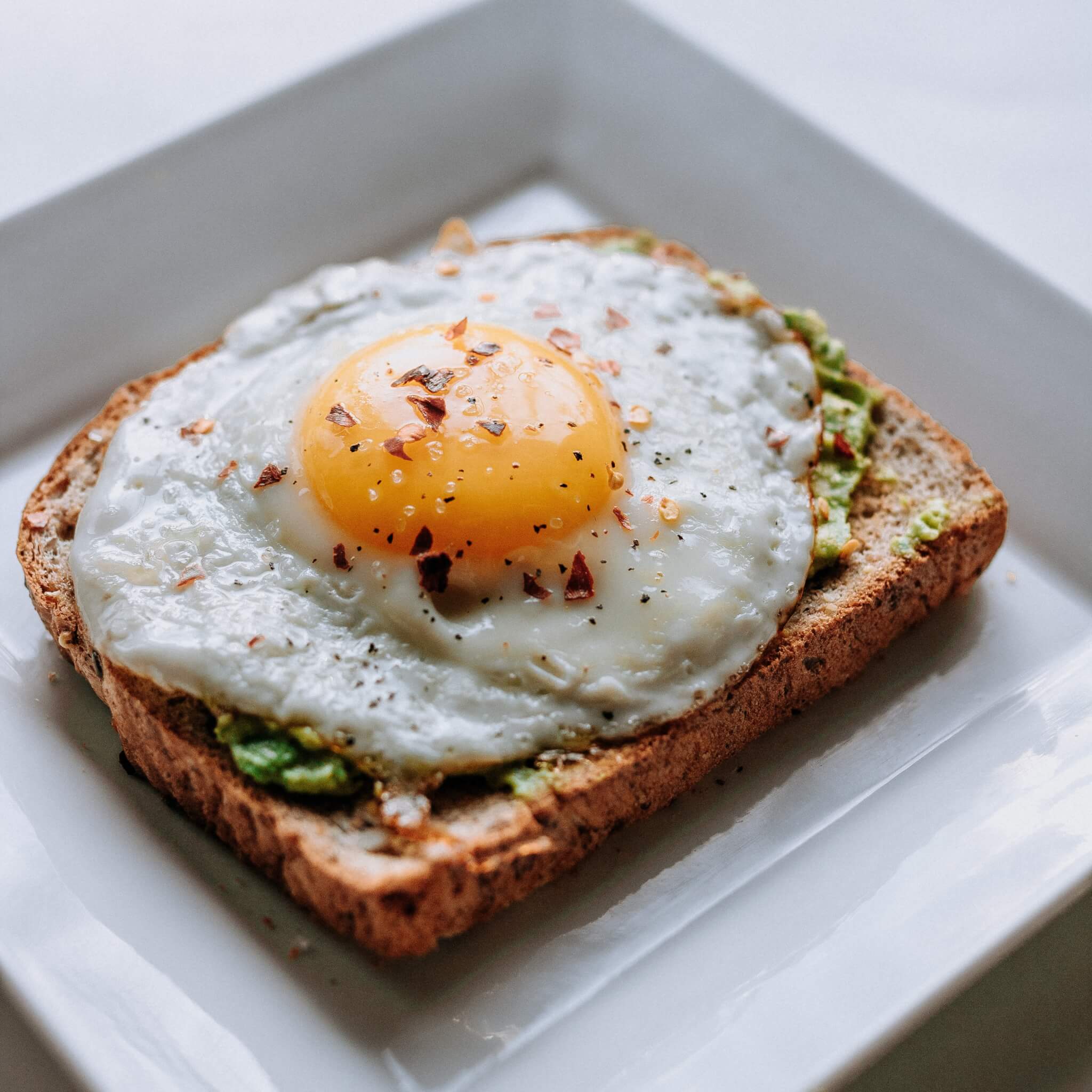 A slice of toast topped with mashed avocado and a sunny-side-up egg, garnished with pepper flakes on a square white plate. - Home Instead