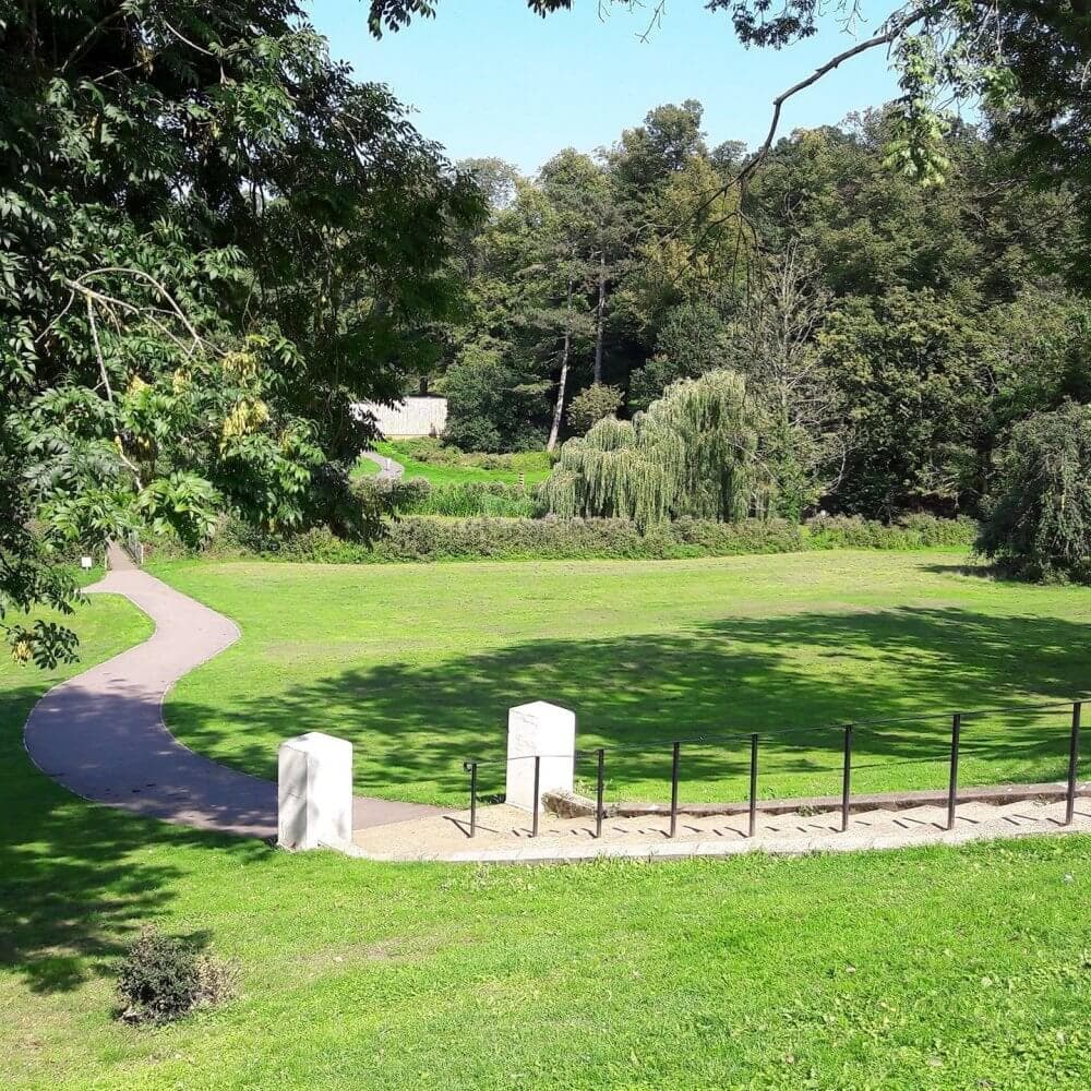 A winding path through a green park with trees and two white stone markers in the foreground on a sunny day. - Home Instead