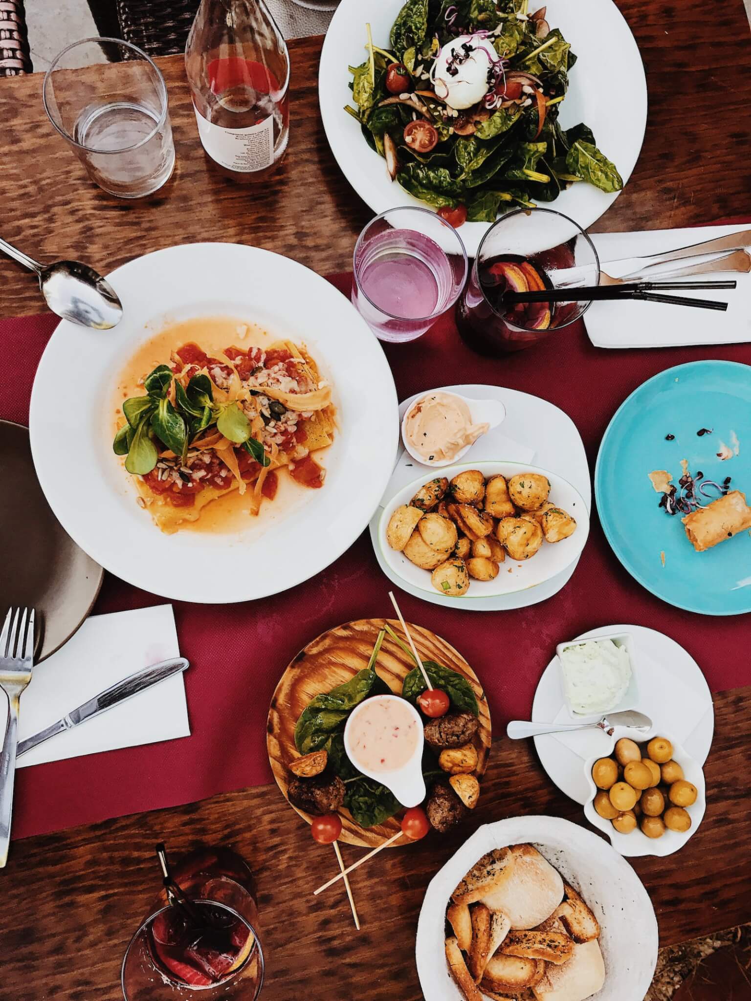 A table set with various gourmet dishes including salads, potatoes, bread, olives, and drinks. - Home Instead