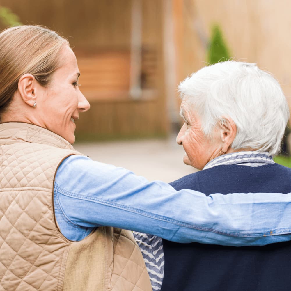 A young woman with a ponytail affectionately wraps her arm around an elderly woman with white hair outdoors. - Home Instead