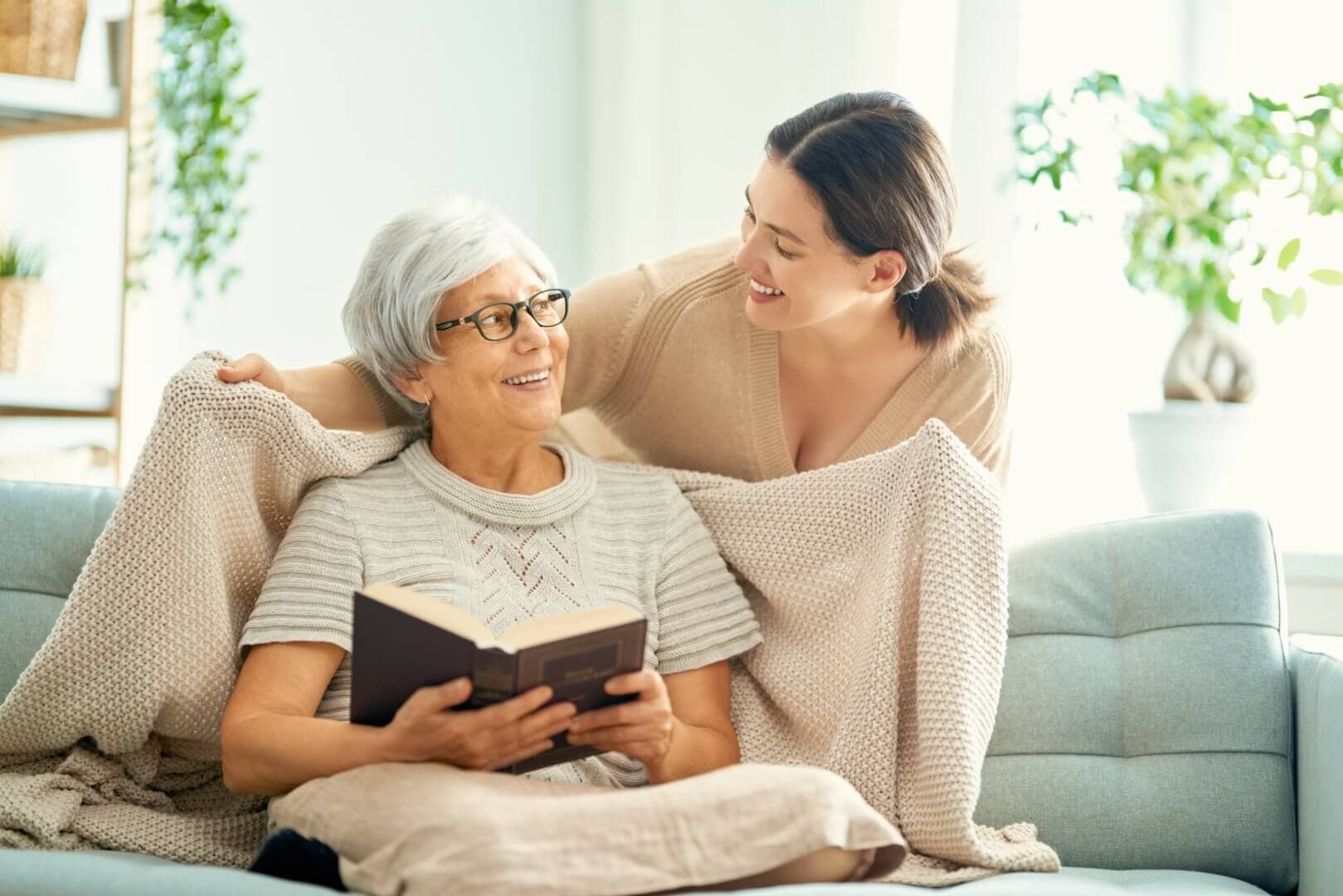 An elderly woman with a book sits on a couch, smiling at a young woman draping a blanket over her shoulders. - Home Instead Bournemouth & Christchurch