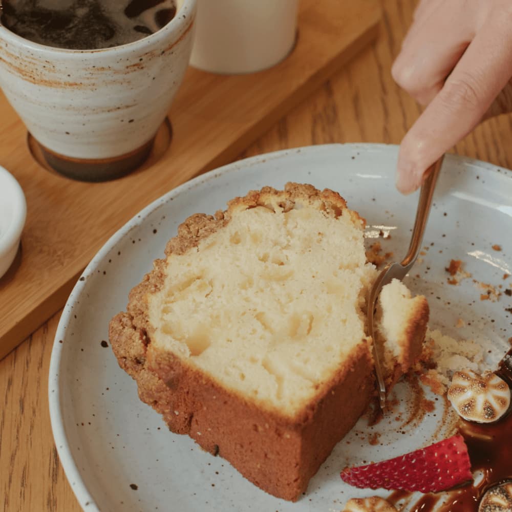 A hand cutting a slice of cake with a fork beside a cup of coffee and a plate with strawberries and chocolate drizzle. - Home Instead