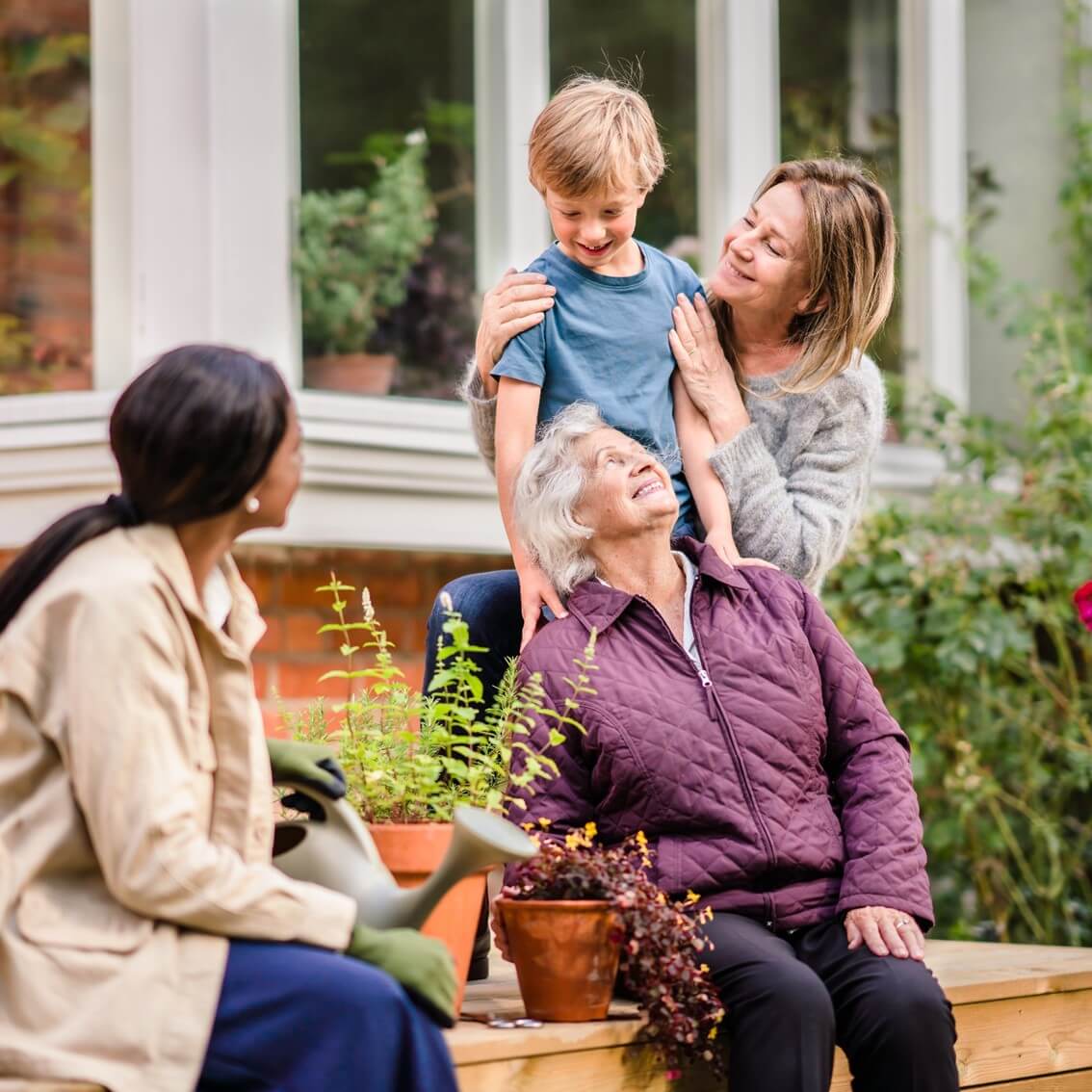 Three generations enjoying time together in a garden, with a child, mother, grandmother, and another woman with a watering can. - Home Instead