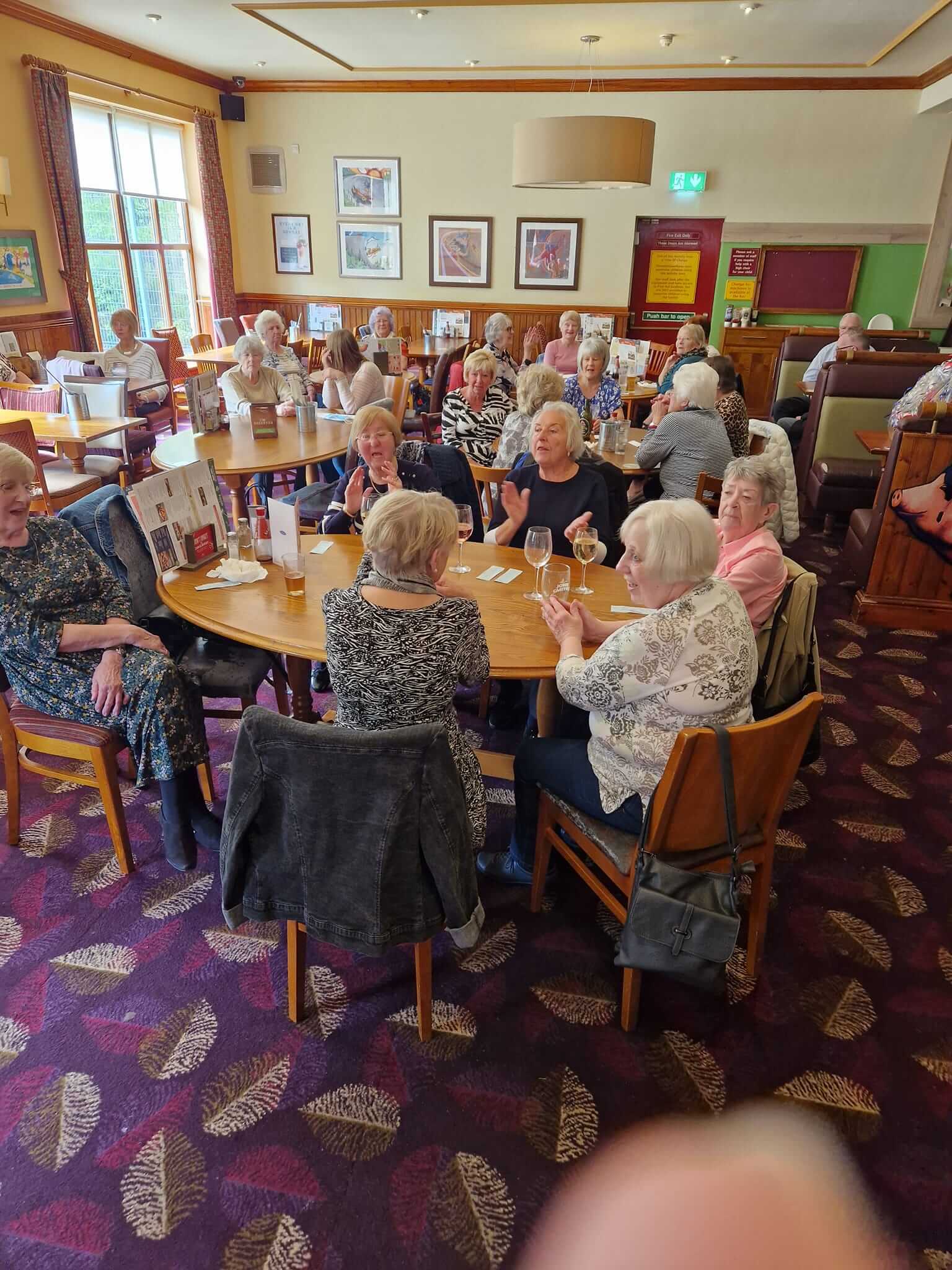 A group of elderly people socializing around tables in a cozy restaurant with patterned carpet and framed pictures on the walls. - Home Instead