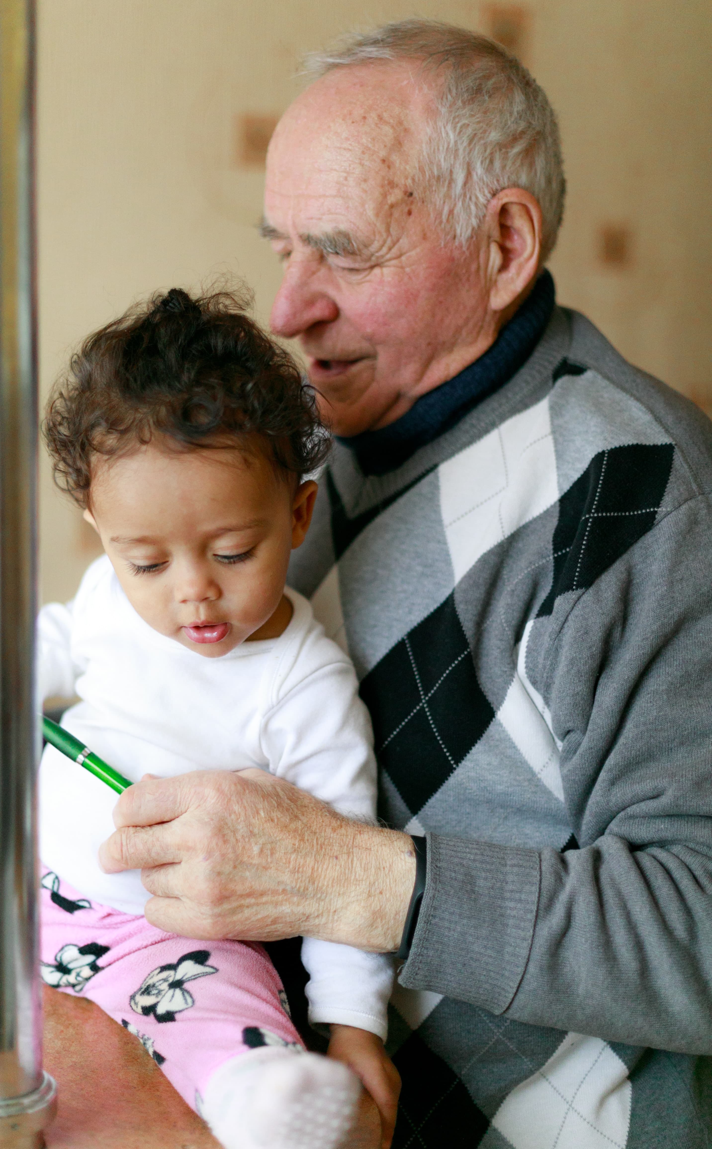 An elderly man holding a toddler on his lap, with the toddler looking at a green object in his hand. - Home Instead