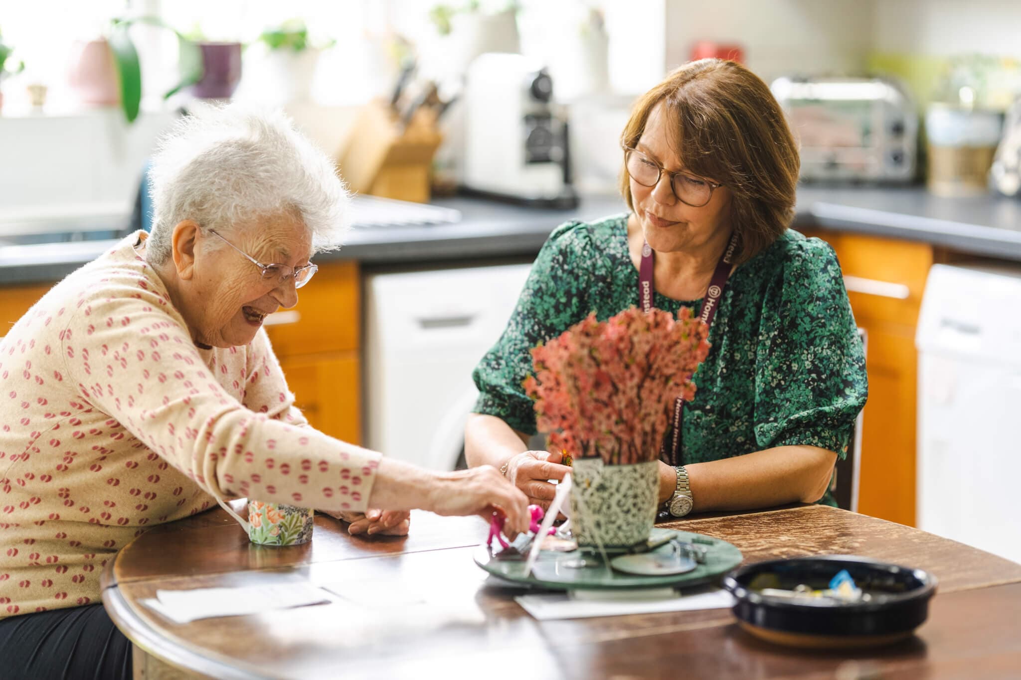 An elderly woman and a caregiver are sitting at a table, engaging in an activity together in a homey kitchen setting. - Home Instead