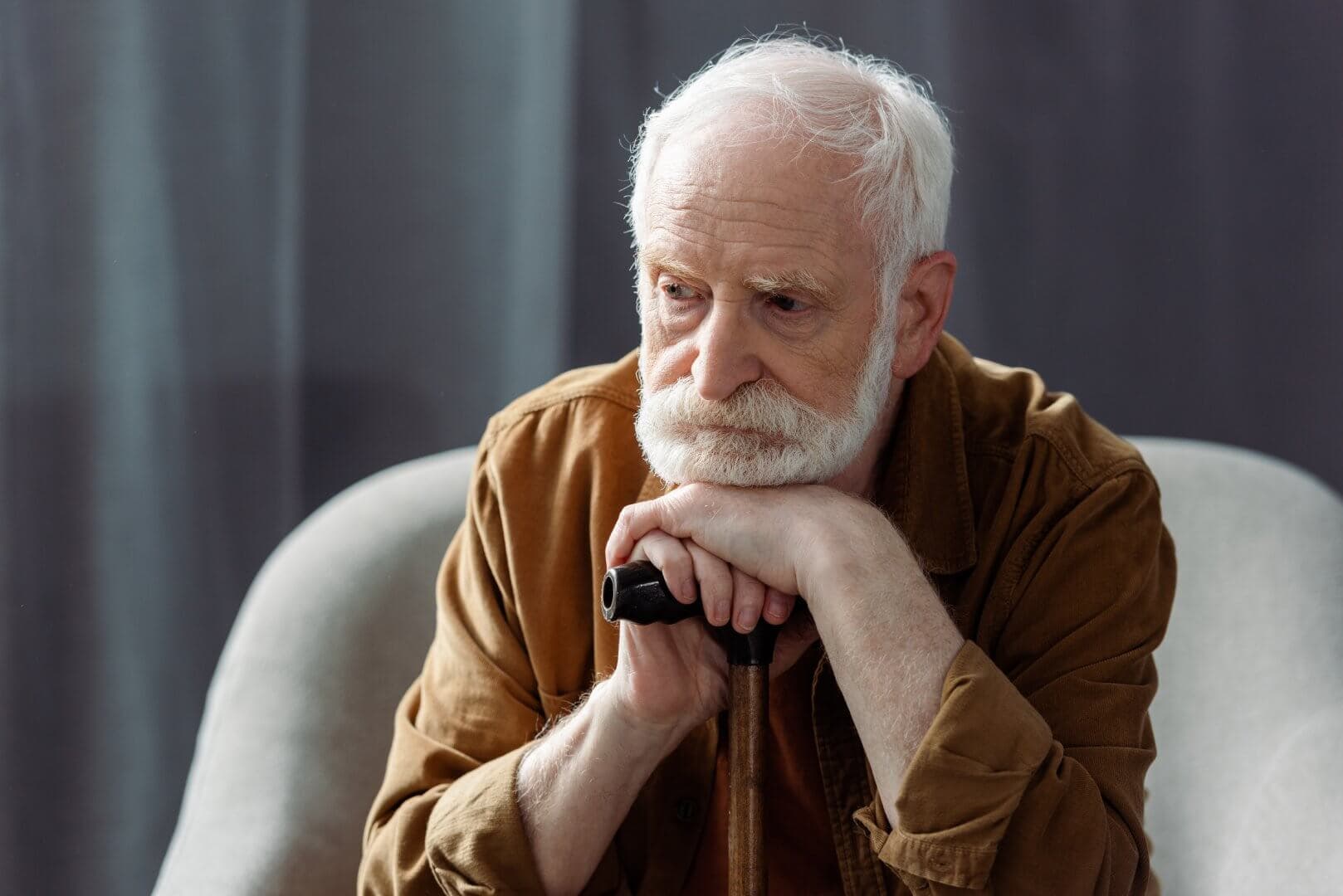Elderly man with white beard sitting thoughtfully, resting his chin on his hands placed on a walking cane. - Home Instead