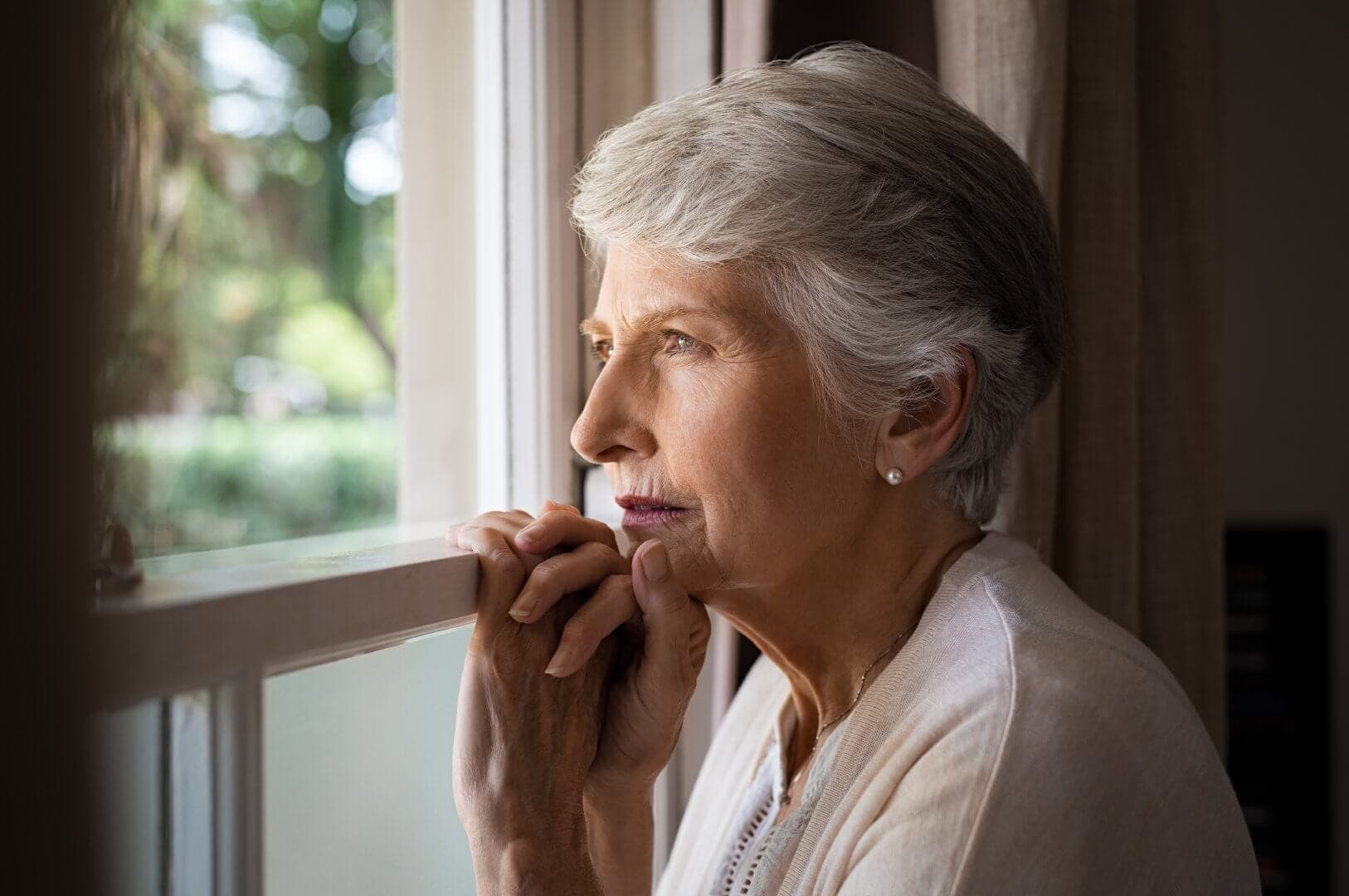 An elderly woman with short grey hair looks pensively out of a window, holding onto the window frame. - Home Instead Bournemouth & Christchurch