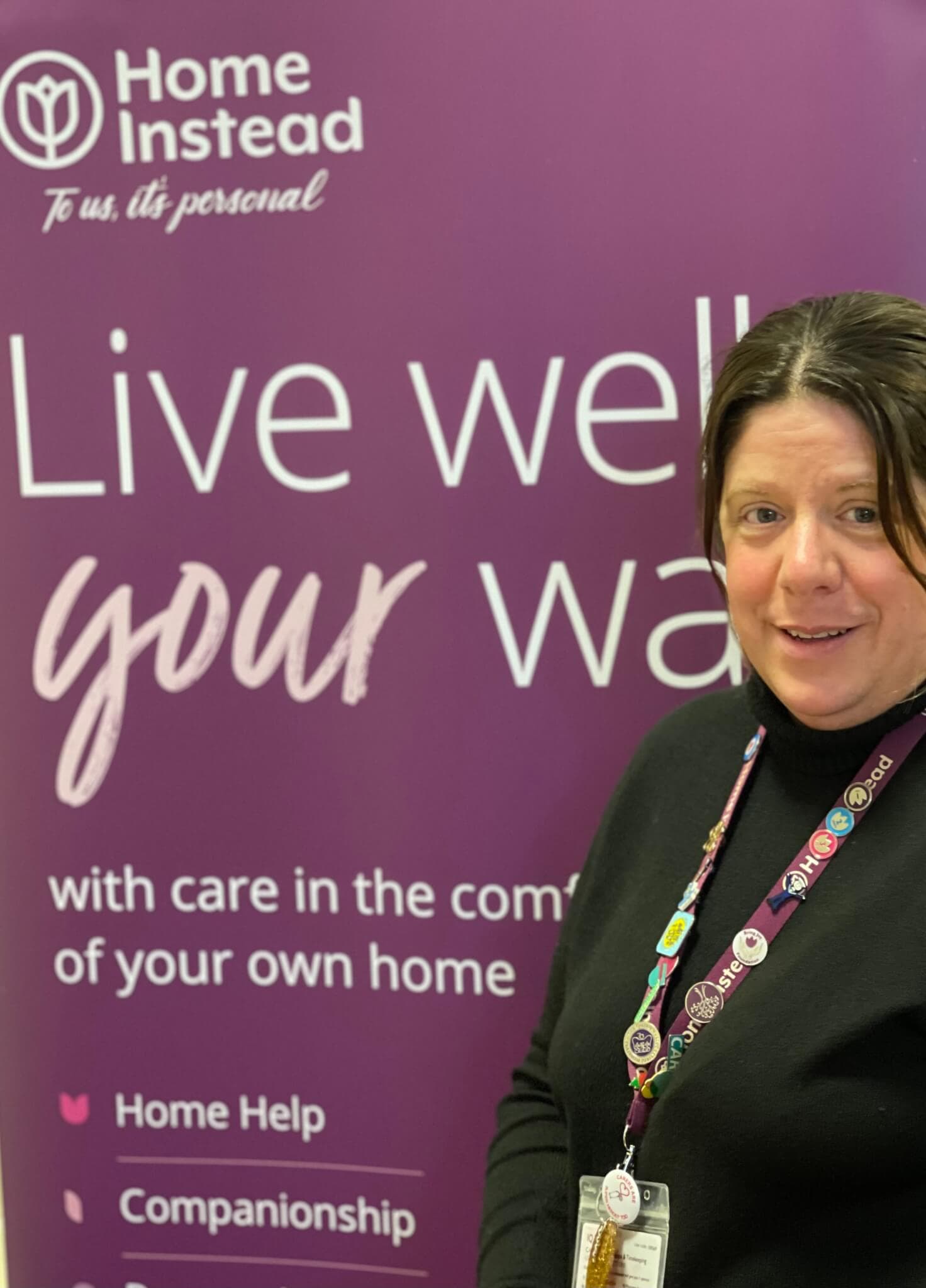 A woman stands in front of a purple "Home Instead" banner that says "Live well your way" with services listed below. - Home Instead
