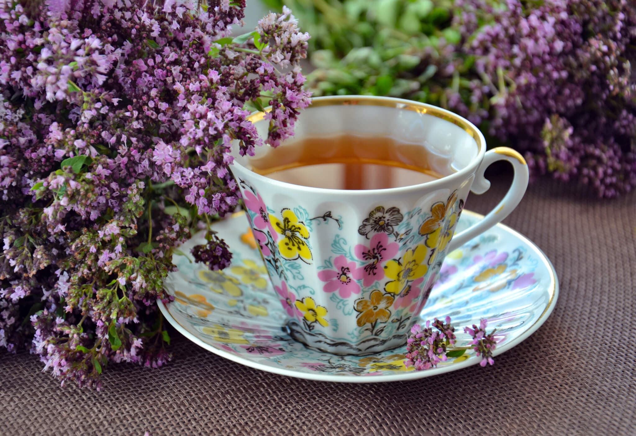 Floral teacup with tea on a matching saucer, surrounded by clusters of purple flowers. - Home Instead
