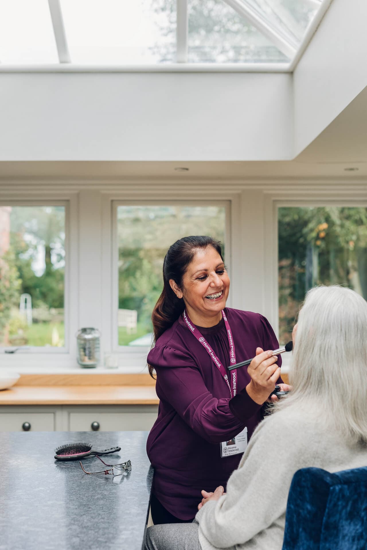 A caregiver with a name tag applies makeup to a seated elderly woman in a bright kitchen with large windows. - Home Instead