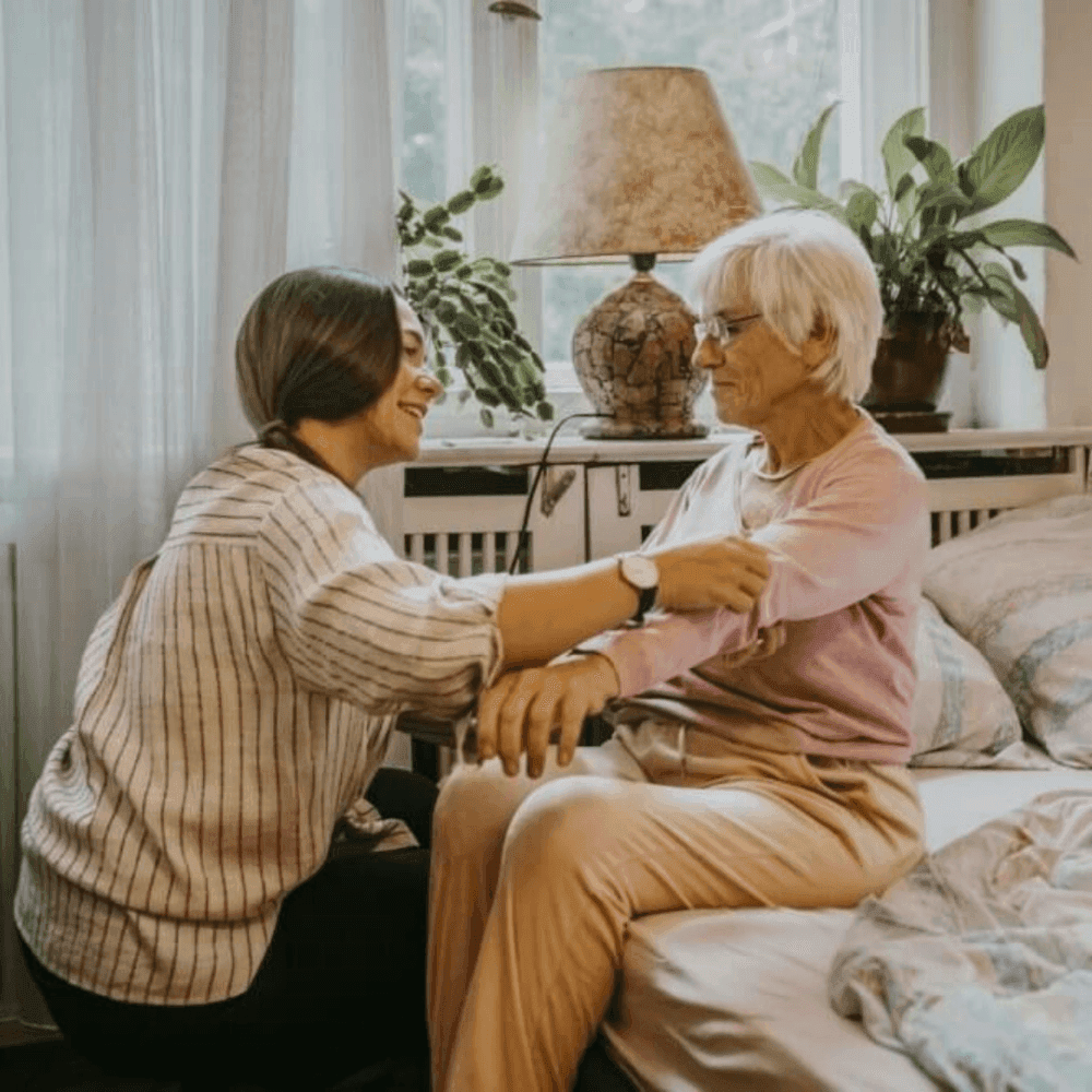 A young woman helps an elderly woman in a bedroom, both smiling, with plants and a lamp visible in the background. - Home Instead