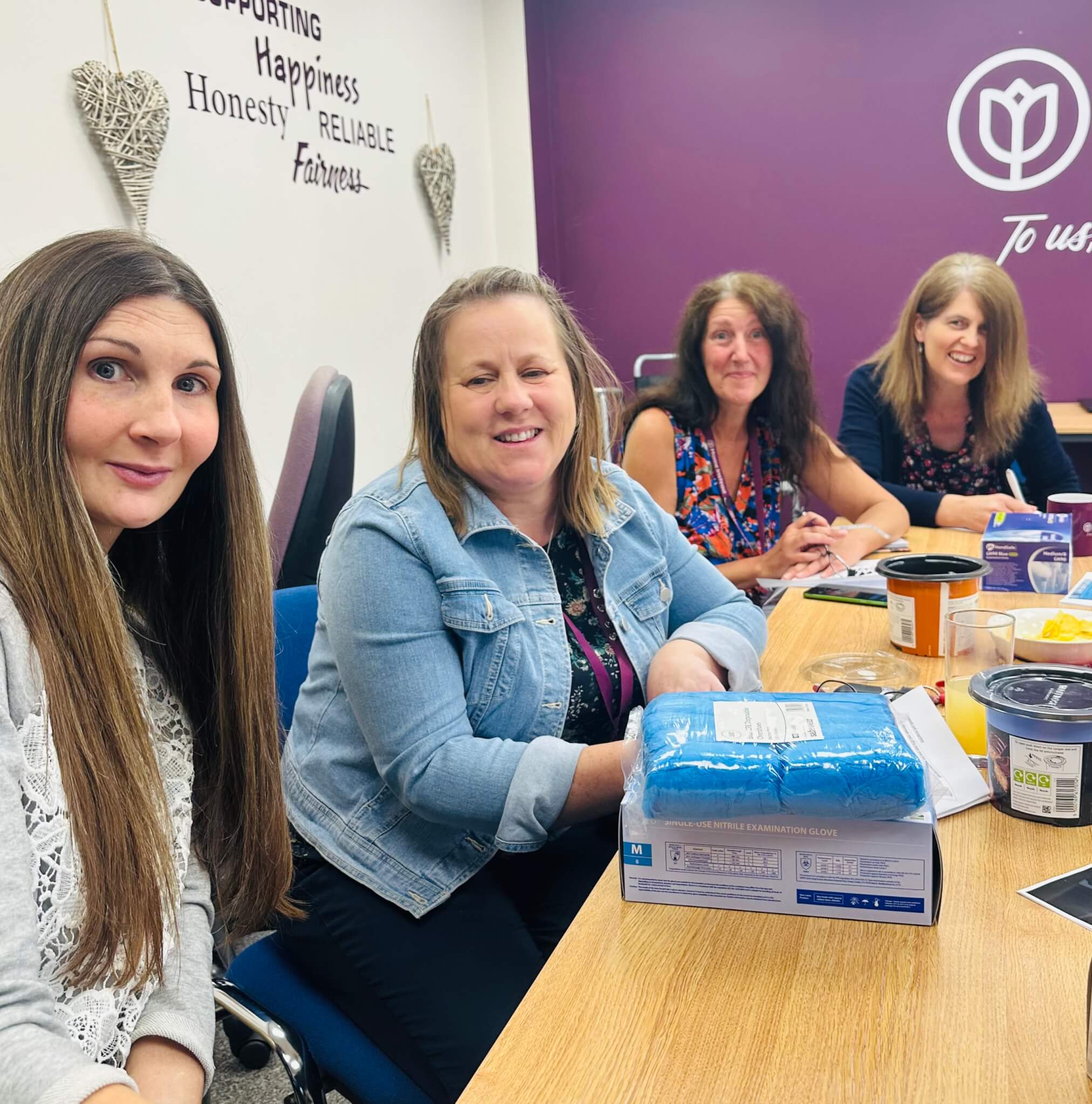 Four women sit around a table, smiling, with various items on the table in a room with a motivational sign on the wall. - Home Instead