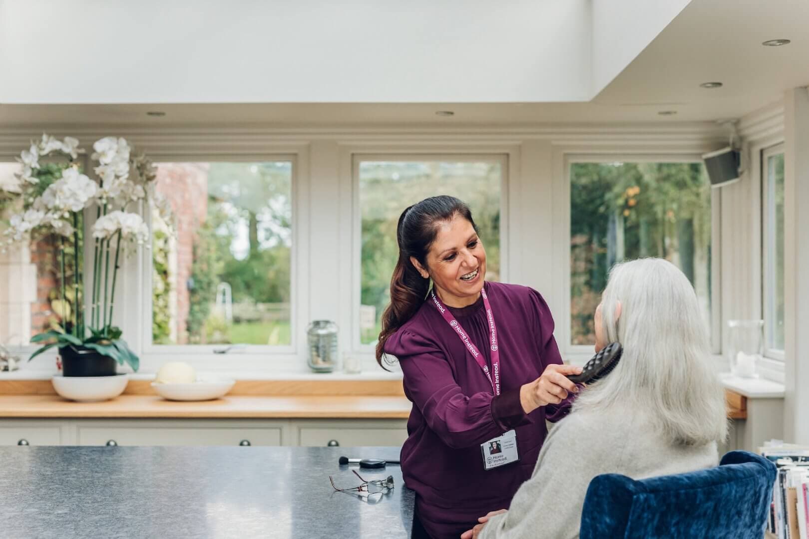 A caregiver in a purple uniform brushes the hair of a seated elderly woman in a bright room with large windows. - Home Instead