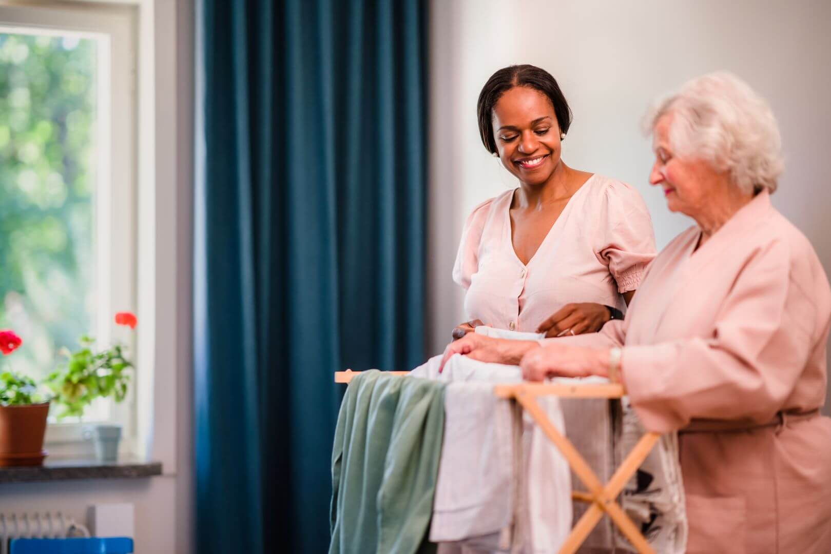 Senior woman and younger woman smiling while folding laundry together inside a bright room. - Home Instead