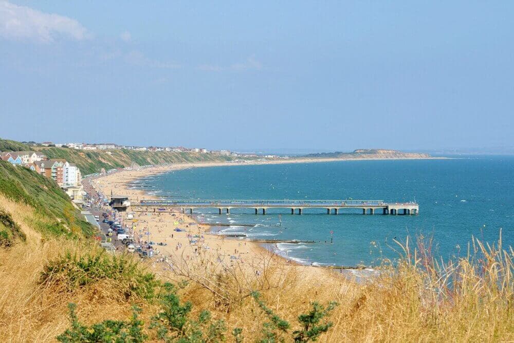 A sandy beach with a pier extends along the coast, with grassy hills in the foreground and buildings in the background. - Home Instead Bournemouth & Christchurch