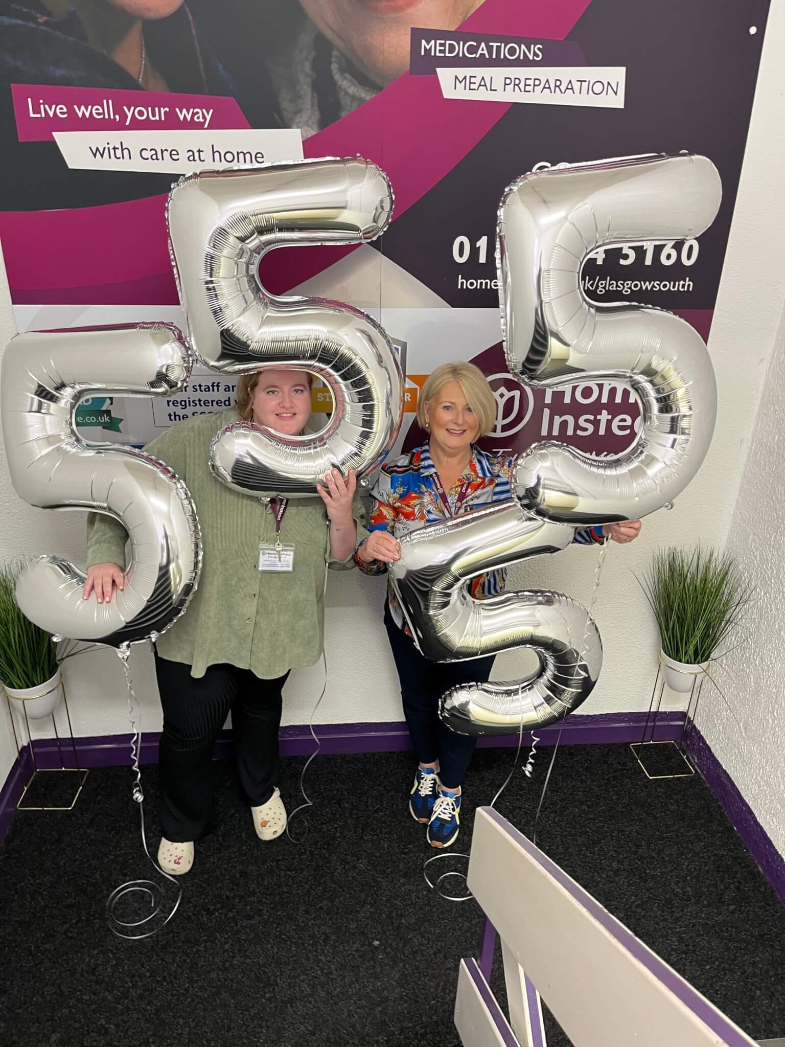 Two women standing and smiling while holding large balloon numbers "555" indoors in front of a branded wall. - Home Instead