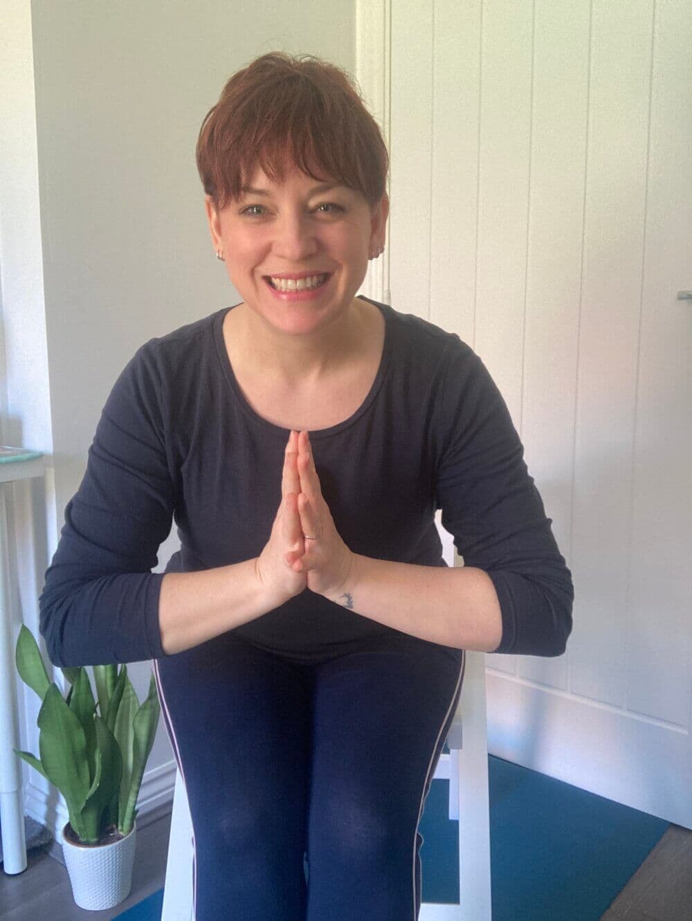 Person smiling and doing a yoga 'Namaste' gesture, sitting on a chair indoors, with a potted plant in the background. - Home Instead