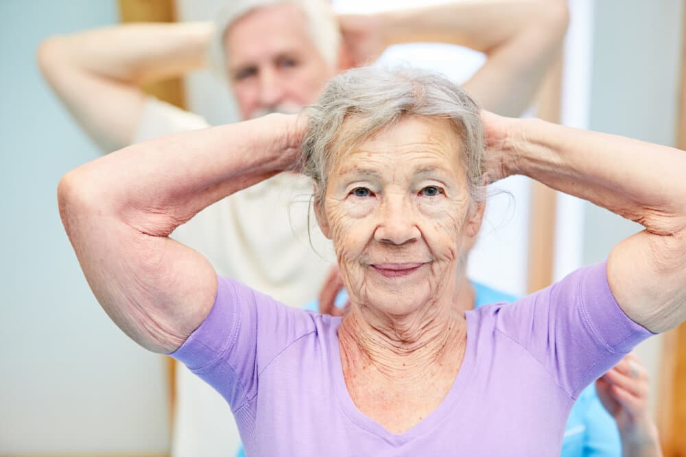 Older woman in a purple shirt exercising with hands behind her head, smiling with people in the background. - Home Instead