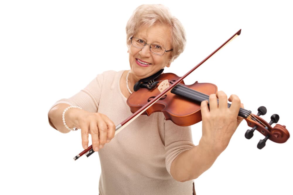 Elderly woman with glasses smiling while playing a violin, wearing a light-colored sweater, isolated on a white background. - Home Instead