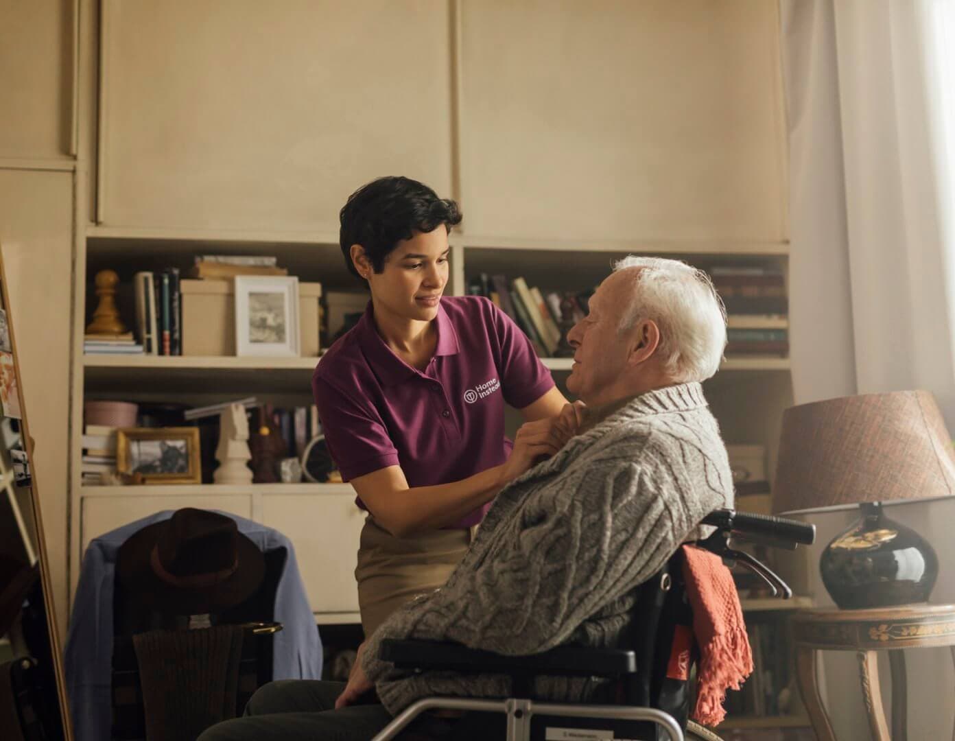 Caregiver in a purple shirt helps an older man in a wheelchair with his scarf in a cozy, book-filled living room. - Home Instead