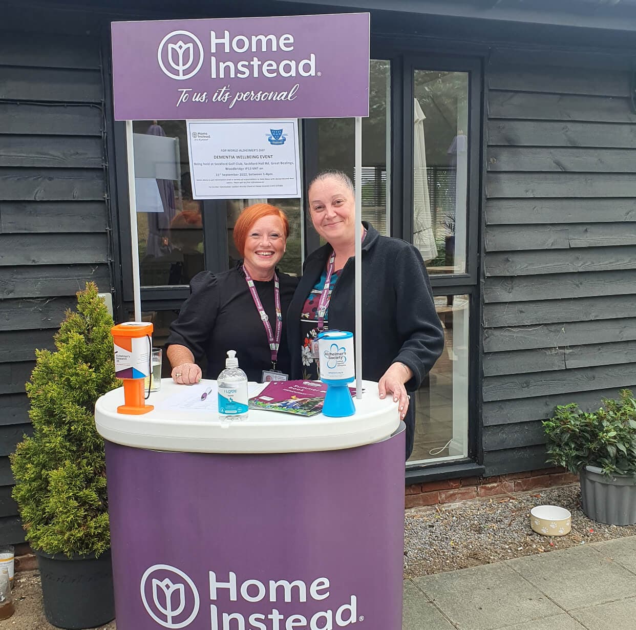 Two women stand at a Home Instead booth with brochures, donation boxes, and a sanitizer bottle on display. - Home Instead