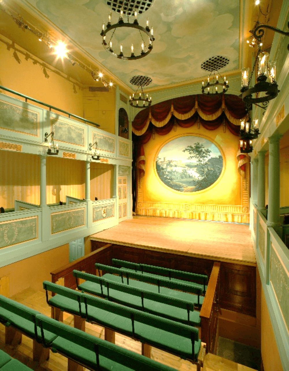 An empty, small vintage theater with green seats, balconies, chandeliers, and a painted stage backdrop. - Home Instead