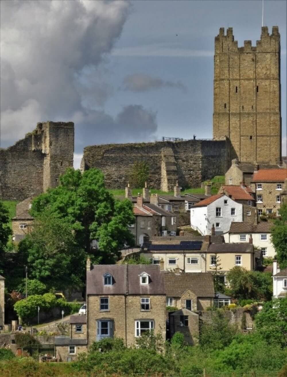 A village with stone houses and a tall medieval castle tower in the background, surrounded by greenery. - Home Instead