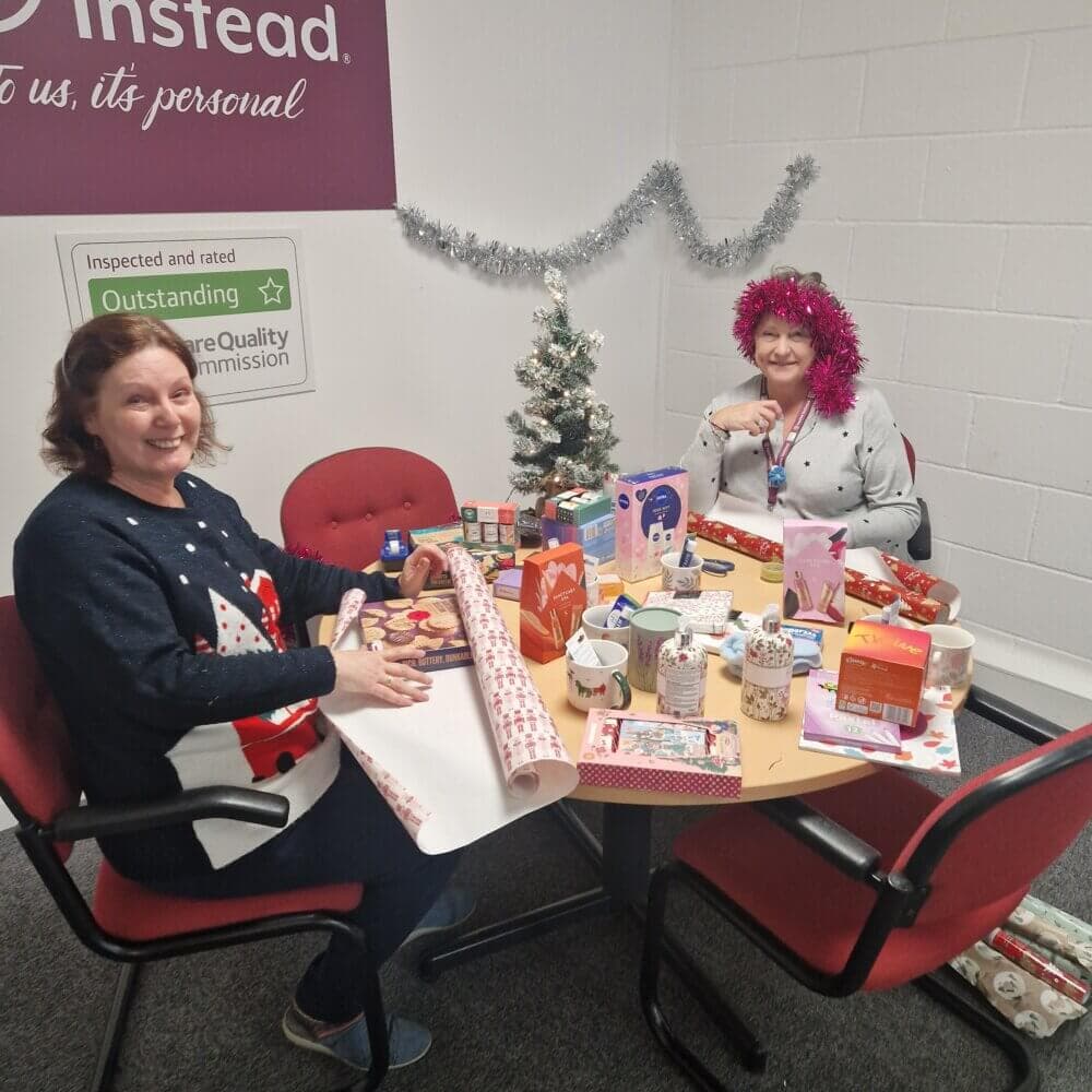 Two women wrapping gifts, sitting at a table with festive decorations and a small Christmas tree in the background. - Home Instead