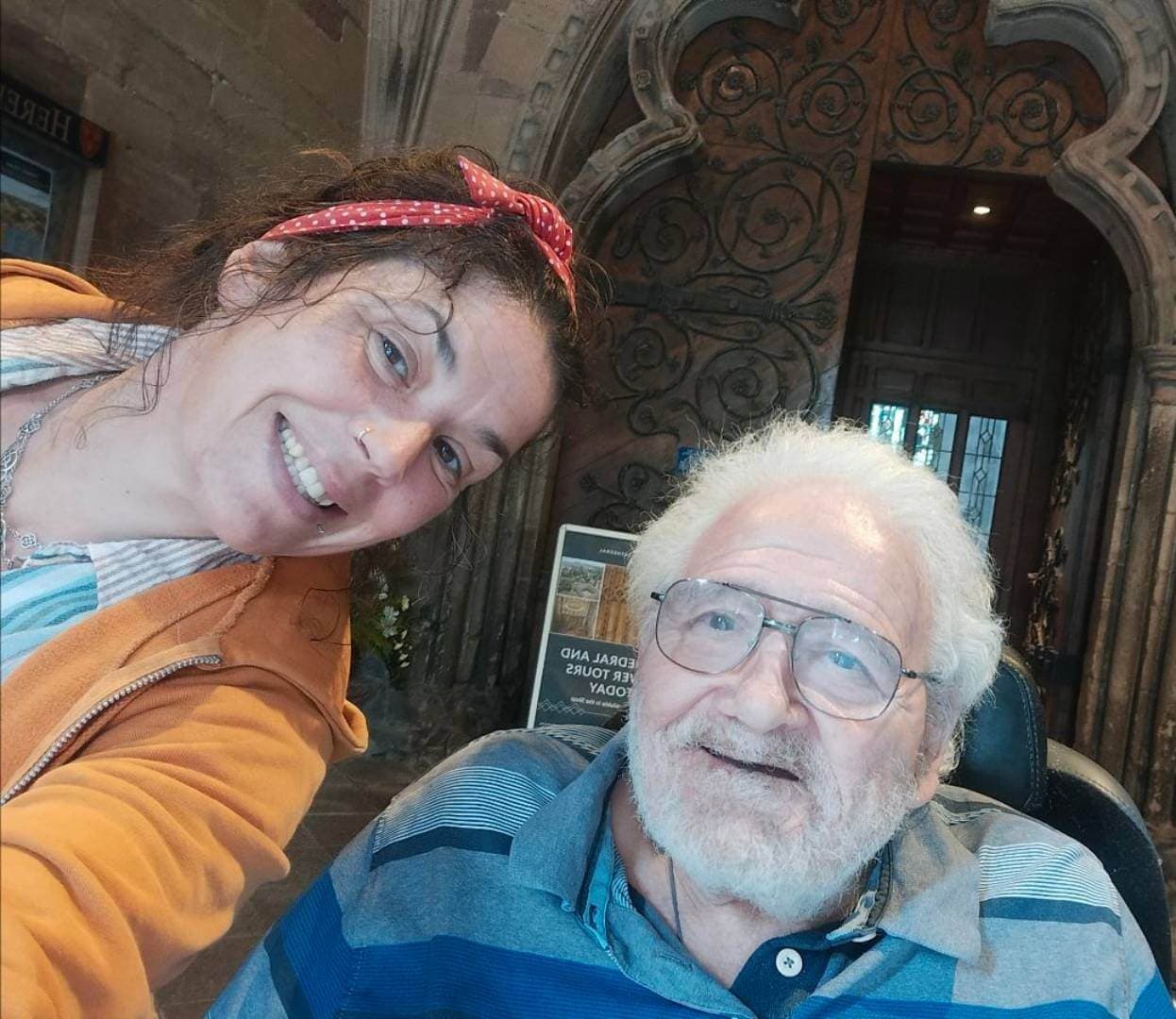 A woman in an orange hoodie and an older man in a wheelchair smile for a selfie inside a building with ornate doors. - Home Instead