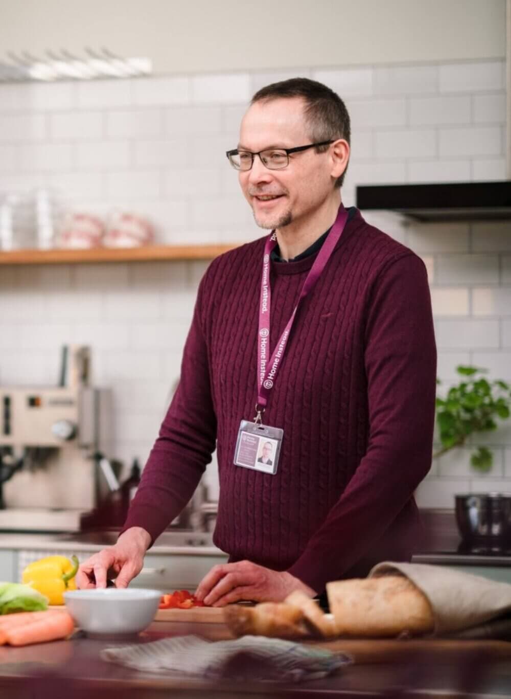A man wearing glasses and a maroon sweater is cutting vegetables in a kitchen, smiling, and wearing an ID badge. - Home Instead