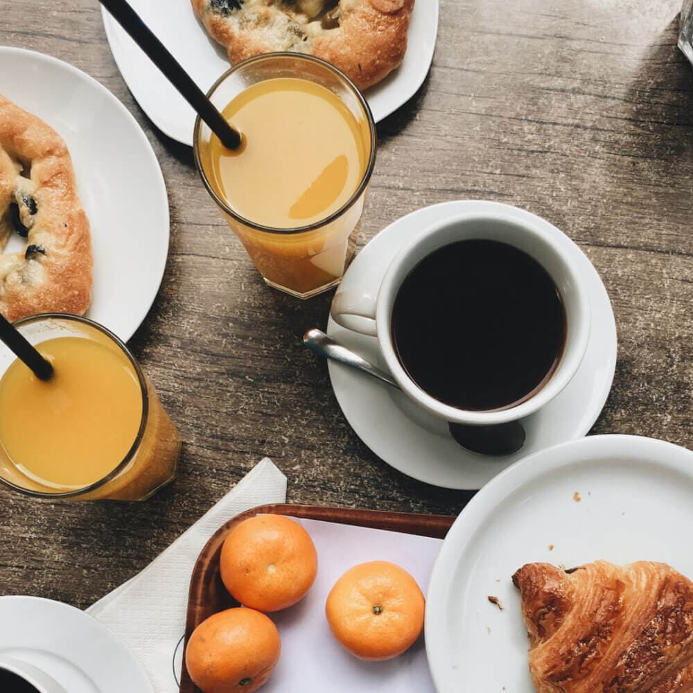Breakfast scene with pastries, croissants, two glasses of orange juice, a cup of coffee, and three small oranges on a table. - Home Instead