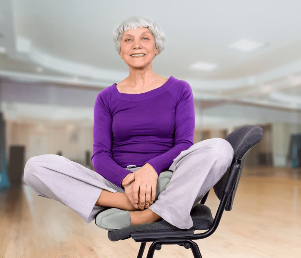 Smiling elderly woman with short white hair in a purple top sitting cross-legged on a chair in a spacious yoga studio. - Home Instead