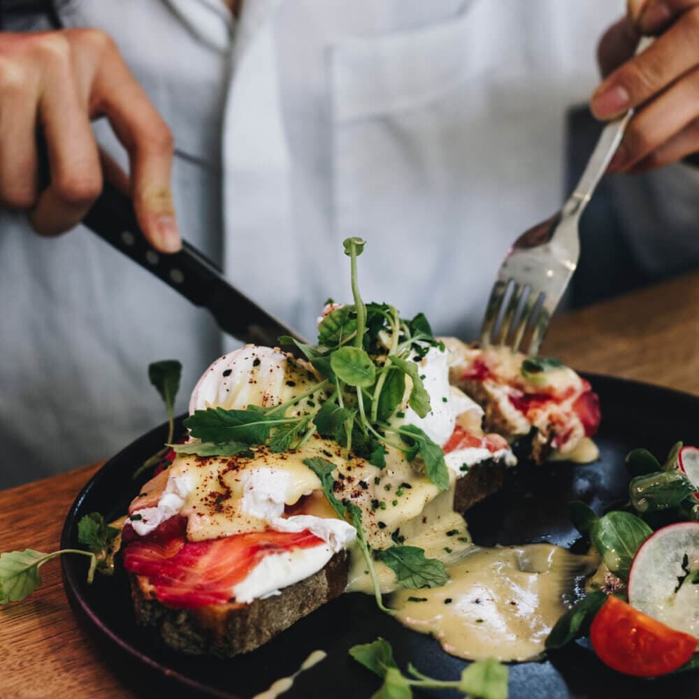 A person cuts into an open-faced sandwich topped with poached eggs, greens, and hollandaise sauce on a black plate. - Home Instead