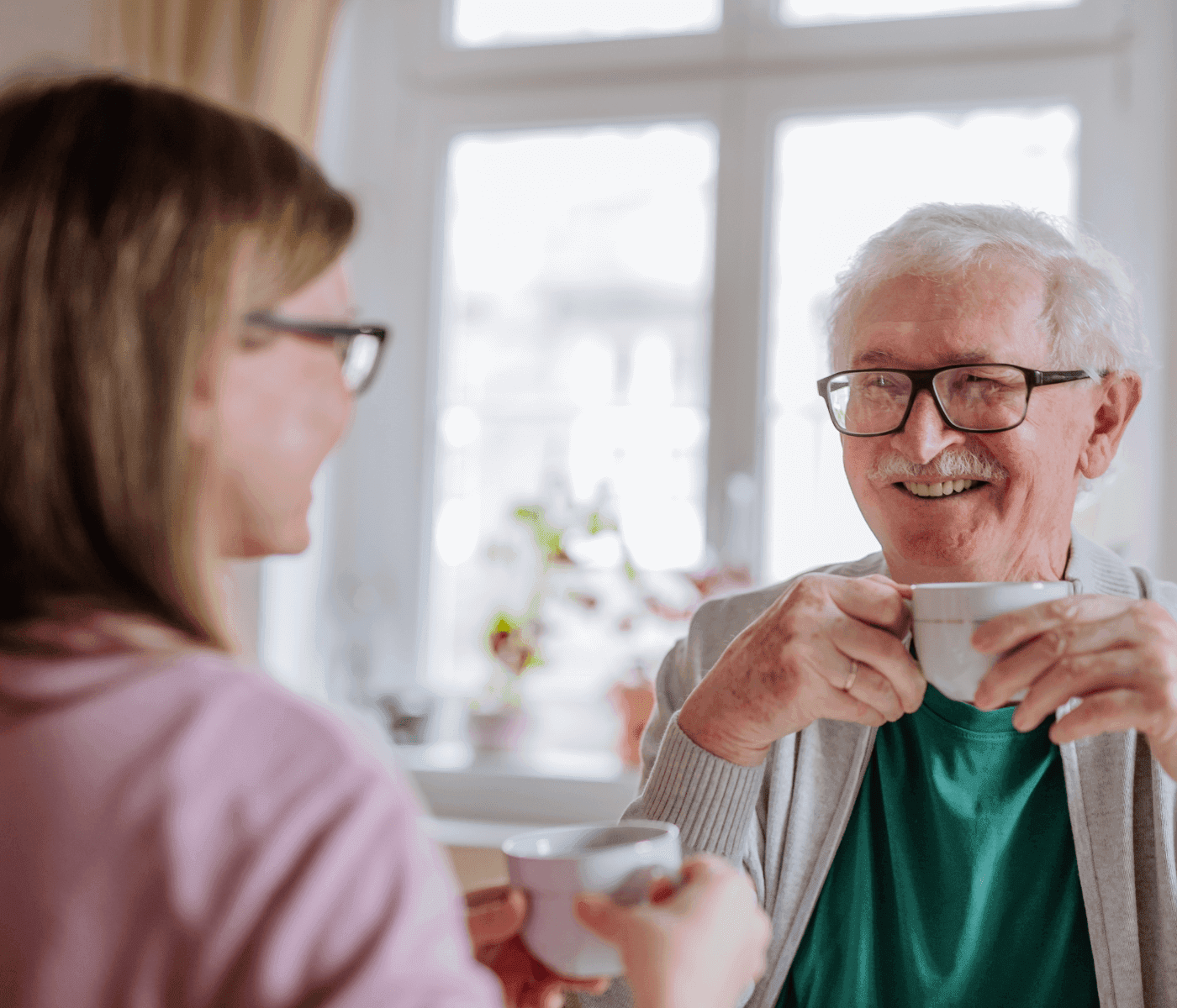 An elderly man and a younger woman smiling and holding coffee mugs while sitting in a brightly lit room. - Home Instead