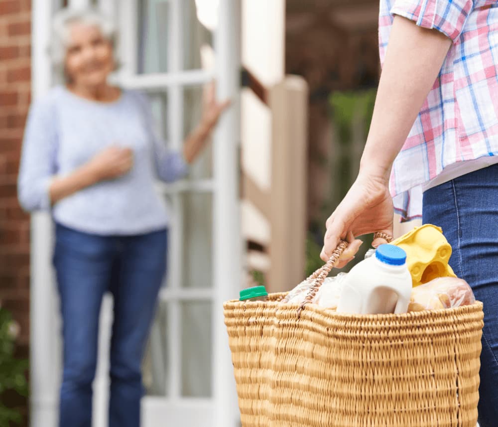 Person delivering a basket of groceries to an elderly individual standing at a door. - Home Instead