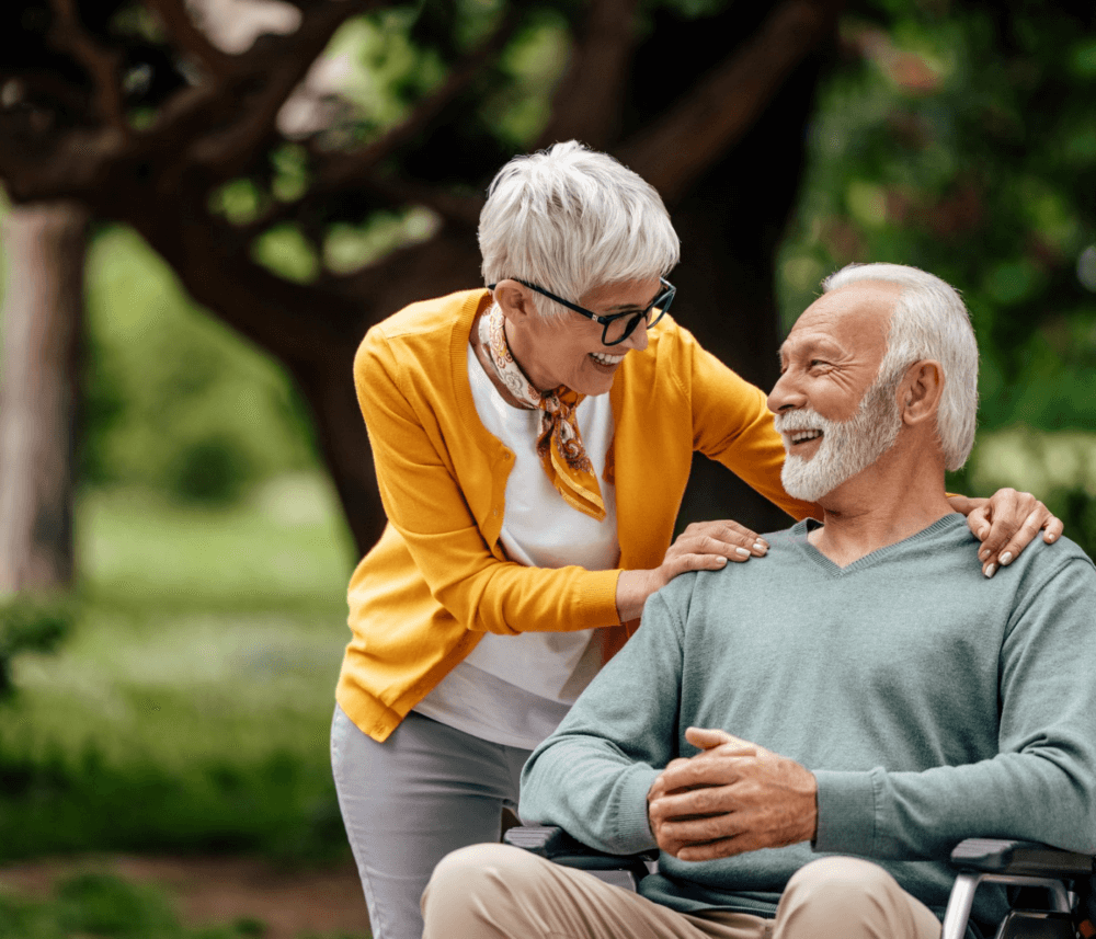 Elderly woman smiles at elderly man in a wheelchair outdoors, both appear happy and engaged in conversation. - Home Instead