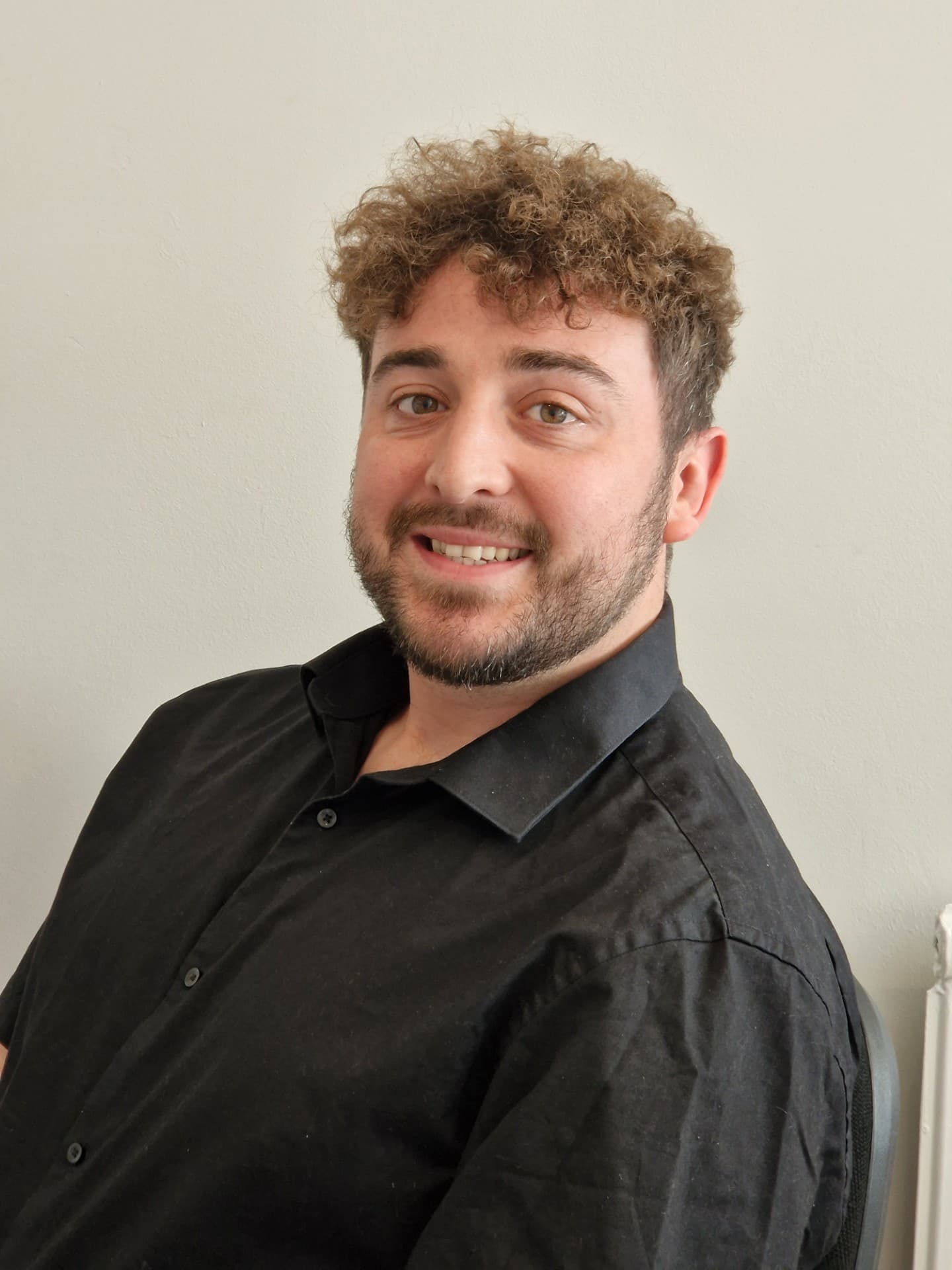 Smiling man with curly hair and beard wearing a black shirt, seated against a plain light-colored wall. - Home Instead