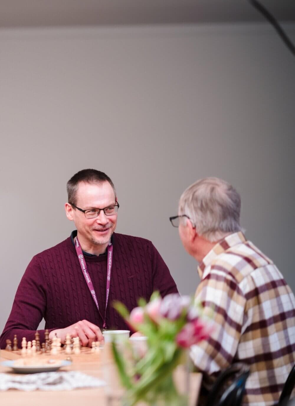 Two men sit at a table with a chessboard, engaged in conversation. One man wears a maroon sweater and a lanyard. - Home Instead