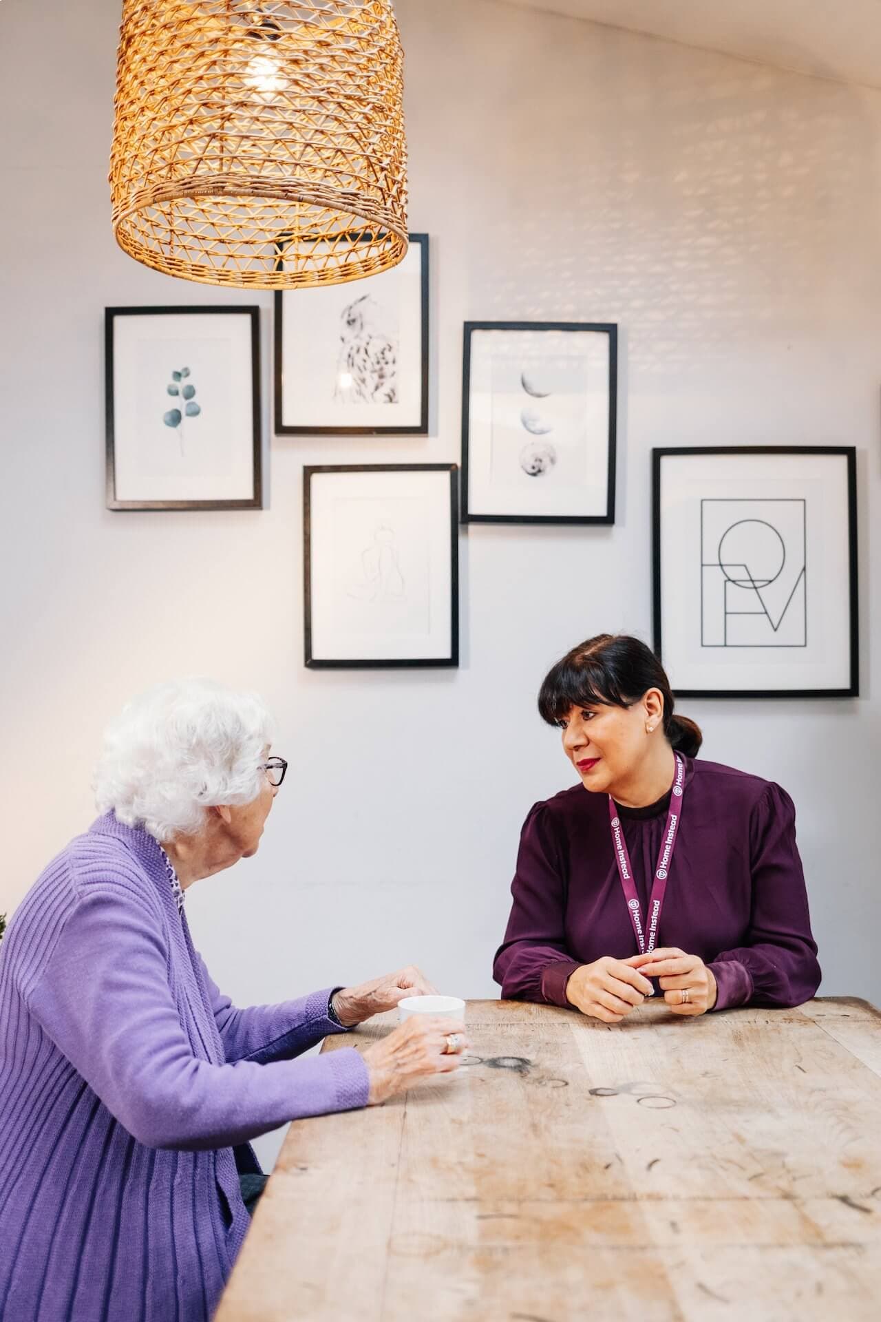 Elderly woman and caregiver having a conversation at a wooden table in a well-lit room with framed art on the wall. - Home Instead
