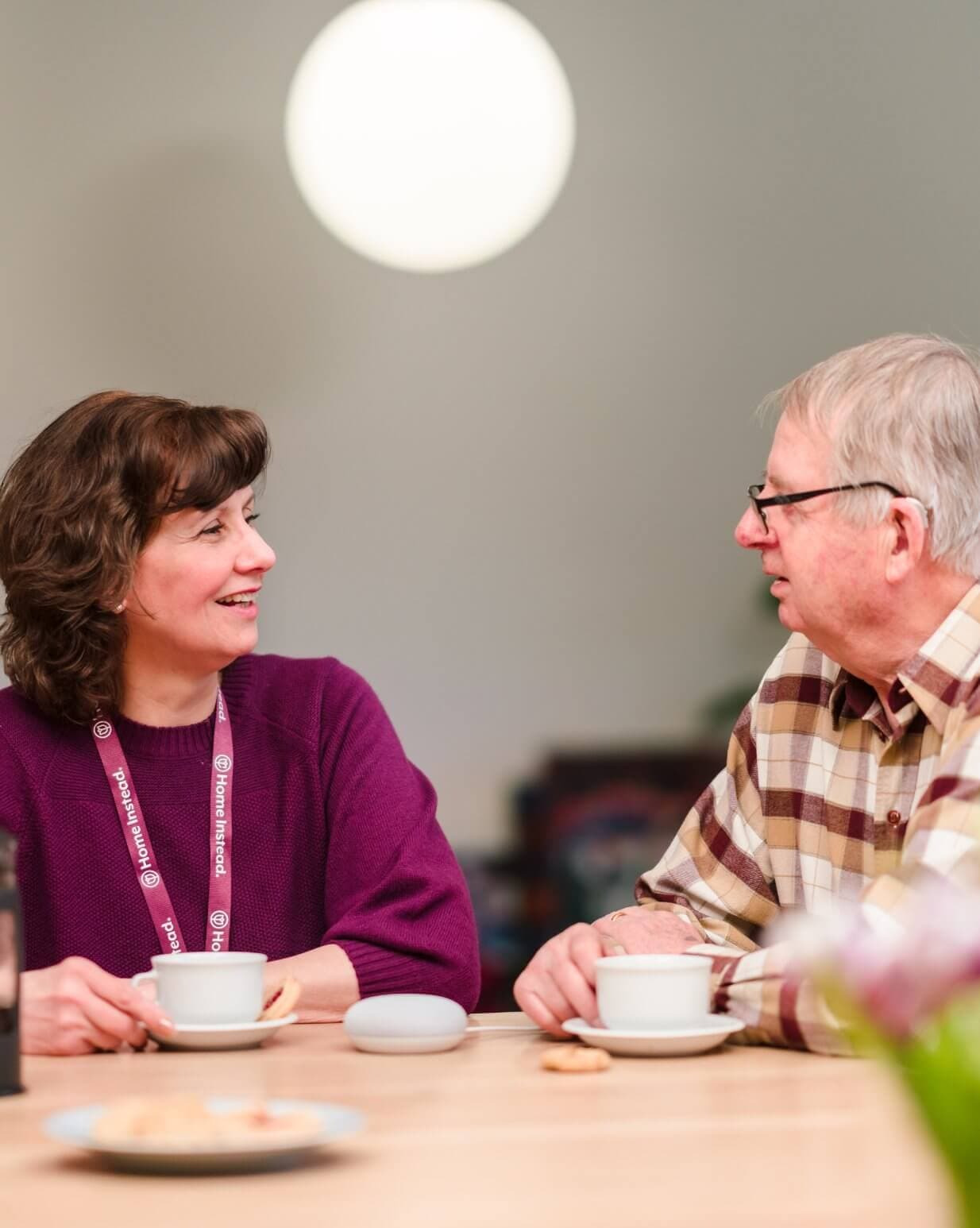 Two people sitting at a table, smiling, sharing a conversation over coffee. - Home Instead