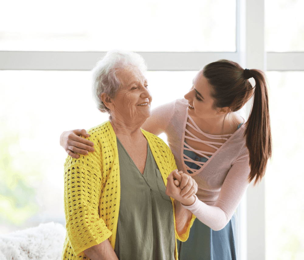 A young woman with a ponytail embraces and smiles at an elderly woman wearing a yellow cardigan in a brightly lit room. - Home Instead