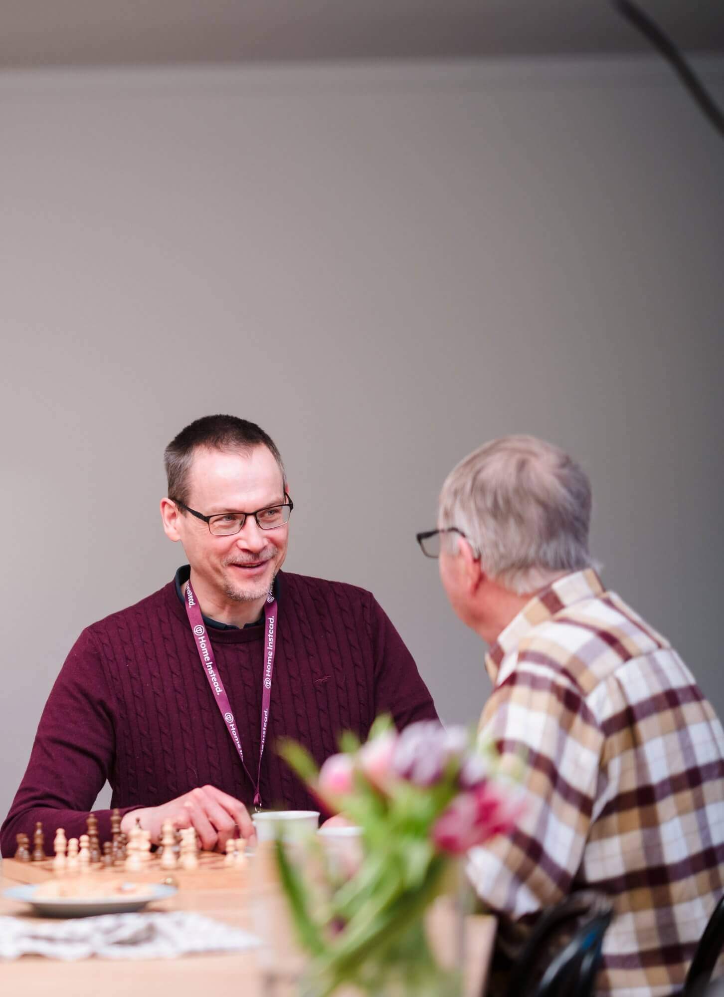 Two men are sitting at a table, smiling and talking, with a chessboard and flowers in the foreground. - Home Instead