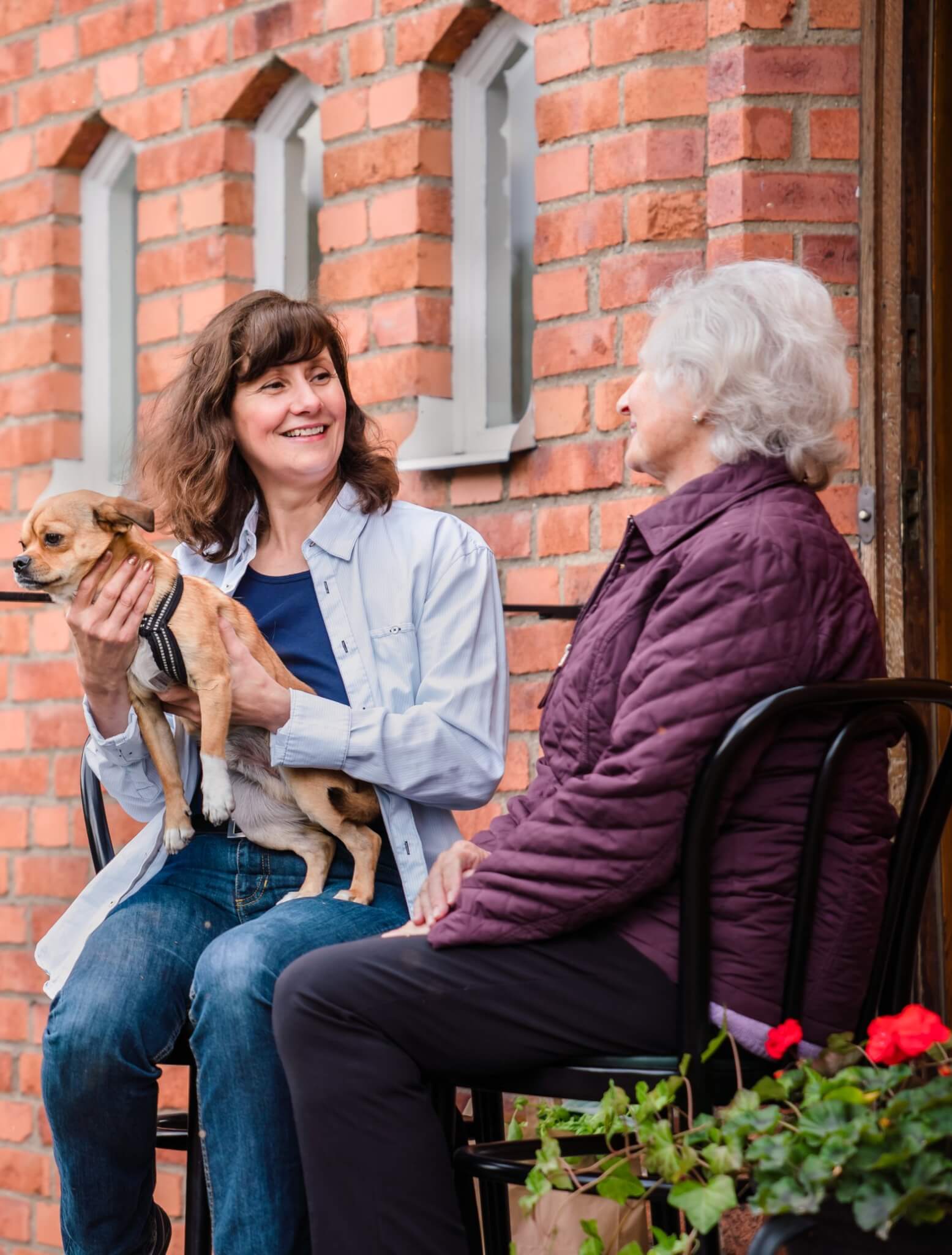 Two women sitting and smiling outdoors, one holding a small dog, with a brick wall in the background. - Home Instead