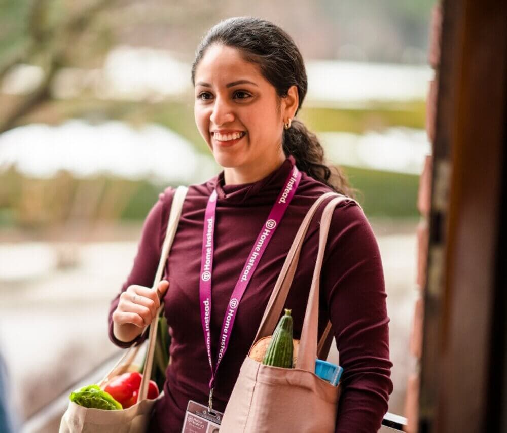 A person smiling and carrying bags of groceries, wearing a maroon shirt and a lanyard with an ID badge. - Home Instead