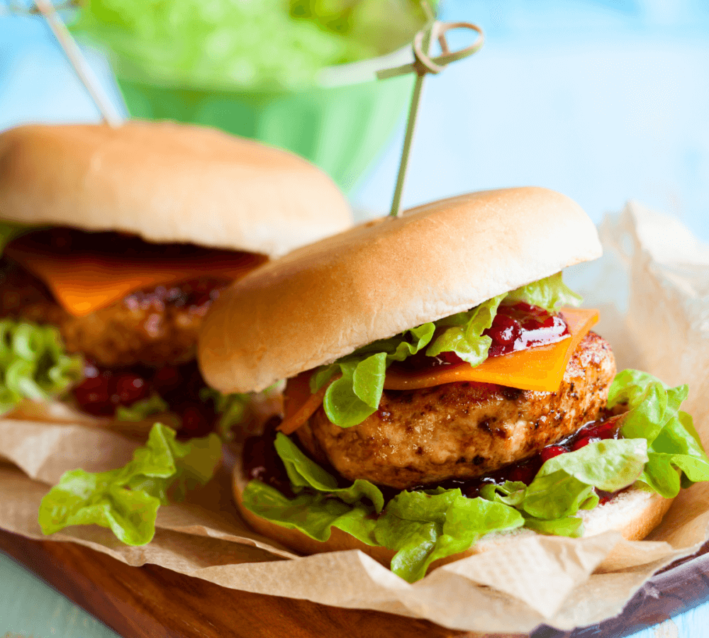 Two juicy chicken burgers with lettuce, cheese, and sauce on a wooden board, garnished with sandwich picks. - Home Instead
