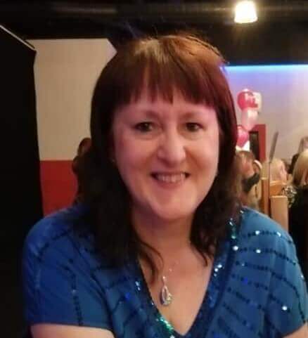 Woman with brown hair and blue top smiling in a dimly lit room, with balloons and people in the background. - Home Instead