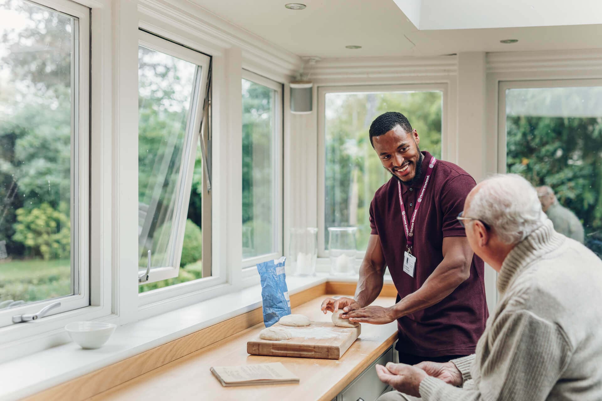 A caregiver and an elderly man share a moment while preparing food in a bright kitchen with large windows overlooking greenery. - Home Instead