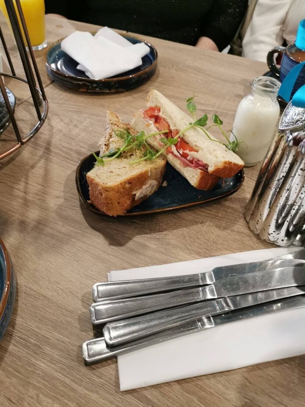 Plate with a sandwich cut in half, garnished with greens, next to cutlery and a small bottle of milk on a table. - Home Instead Renfrewshire and Barrhead home care services - Free monthly client Get Together at Familia Cafe, Renfrew